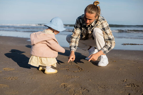 Young dad pointing at heart on sand near baby daughter on beach in Italy 