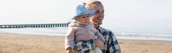 Smiling parent holding toddler kid on beach near adriatic sea in Italy, banner 