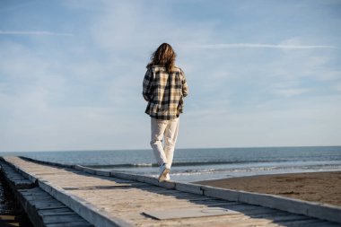 Back view of long haired man walking on wooden pier near adriatic sea in Italy  clipart