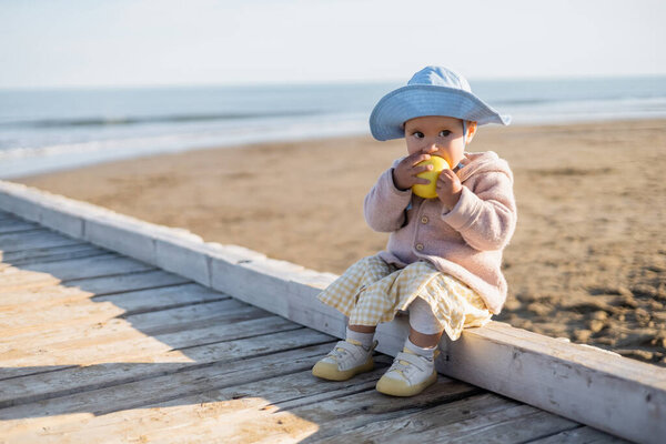 Toddler kid eating ripe apple while sitting on wooden pier in Italy 