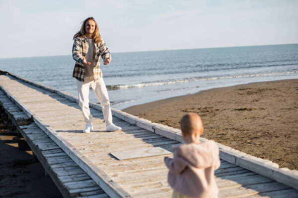 Cheerful man looking at blurred toddler daughter on pier near adriatic sea in Italy 