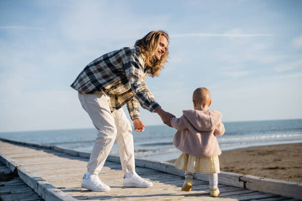 Smiling long haired man holding hand of baby girl on wooden pier near adriatic sea in Italy 