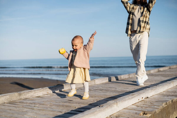 Toddler girl holding apple while walking on pier near father in Italy 