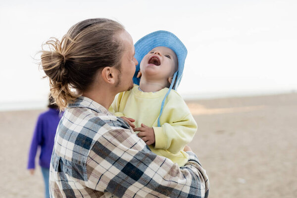 Father holding laughing toddler child in panama hat on beach 