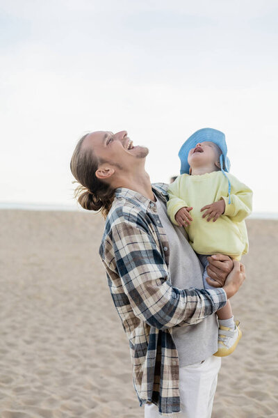 Young man and toddler daughter laughing on beach 