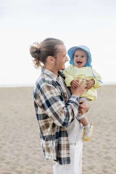 Gülümseyen baba, kumsalda panama şapkalı bebeği tutuyor. 