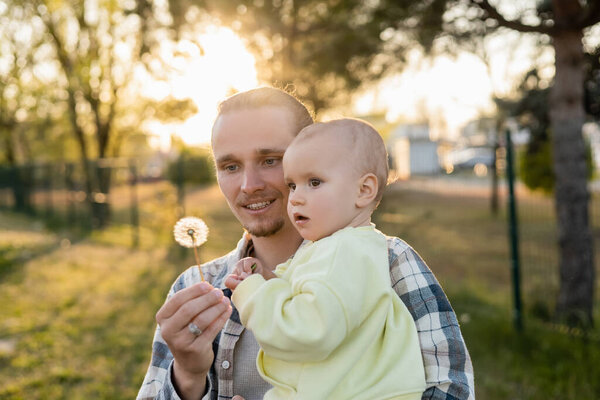 Улыбающийся мужчина держит на улице одуванчика и маленькую дочь 
