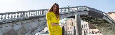 Arka planda Venedik köprüsü yakınlarında poz veren sarı modaya uygun takım elbiseli kızıl saçlı kadın, afiş
