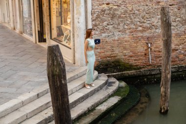 full length of elegant woman on stairs near water and wooden pilings in Venice clipart