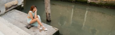 Venedik 'te kanal kenarındaki merdivenlerde oturan genç ve zarif kadının yüksek açılı görüntüsü, afiş.