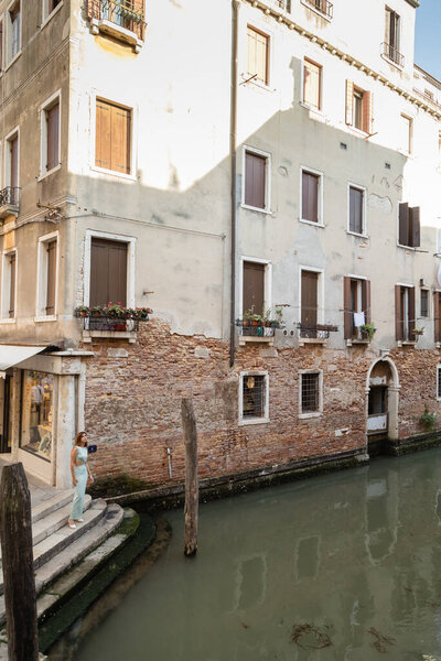 full length of young woman on stairs near medieval building and urban canal in Venice
