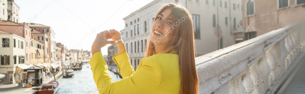 cheerful woman looking at camera while showing heart sign in Venice, banner