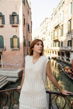 pretty young woman standing near fence over canal in Venice and looking away clipart