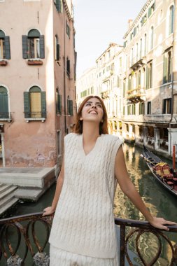 joyful woman looking up while standing near medieval buildings over canal in Venice clipart
