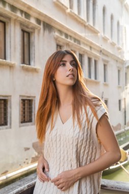 young redhead woman looking away near blurred medieval building in Venice clipart
