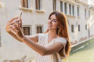Venedik 'teki bulanık binanın yakınındaki akıllı telefondan selfie çeken neşeli kadın.