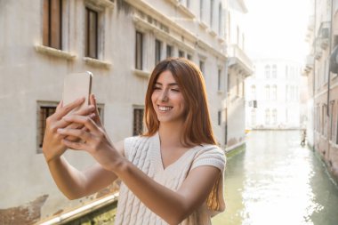 Venedik 'te cep telefonundan selfie çeken kolsuz kazaklı neşeli bir kadın.