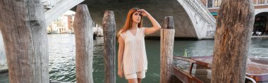 young woman in sleeveless jumper and shorts looking away near wooden pilings in Venice, banner clipart