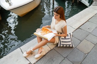 Venedik 'te kaldırımda oturan çizgili çantalı ve lezzetli pizzalı genç bir kadının yüksek açılı görüntüsü