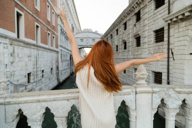 back view of redhead woman with outstretched hands near medieval prison and Sighs Bridge in Venice clipart