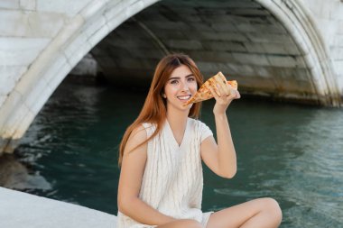 Lezzetli pizzalı neşeli kadın Venedik kanalının yakınındaki kameraya bakıyor.