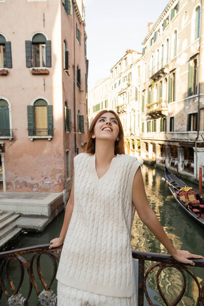 joyful woman looking up while standing near medieval buildings over canal in Venice