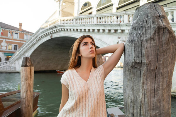 young woman looking away near wooden piling and Rialto Bridge on background in Venice