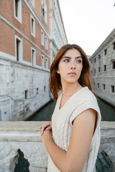 young woman in sleeveless jumper looking away near blurred venetian buildings