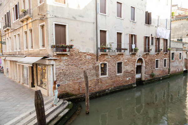 young woman in dress standing near buildings and canal on medieval street in Venice