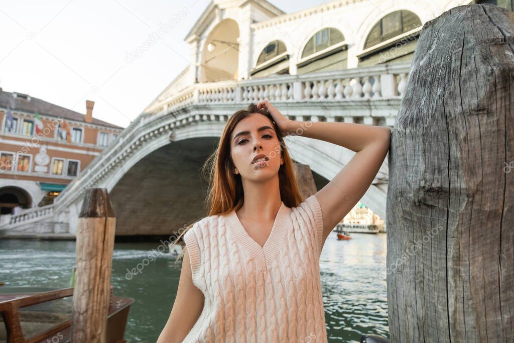 redhead woman looking at camera near wooden piling and Rialto Bridge in Venice