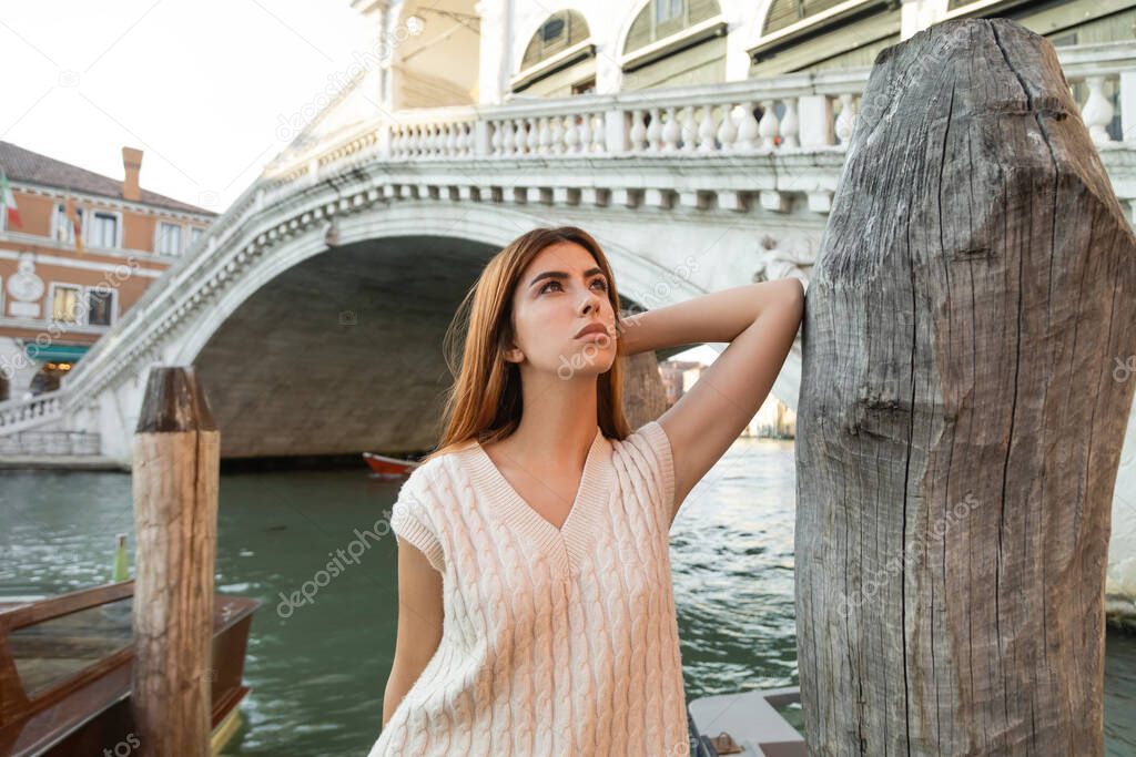young woman looking away near wooden piling and Rialto Bridge on background in Venice