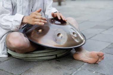 Cropped view of barefoot man playing hang drum on urban street 