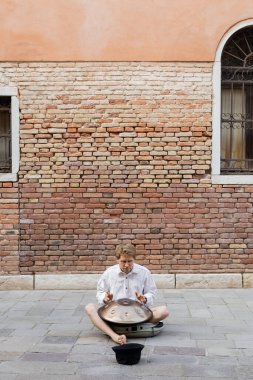 Musician playing hang drum near hat and building on urban street in Venice 