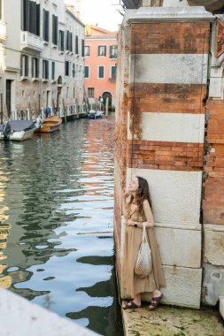Side view of smiling pregnant woman with string bag standing near building and river in Venice 