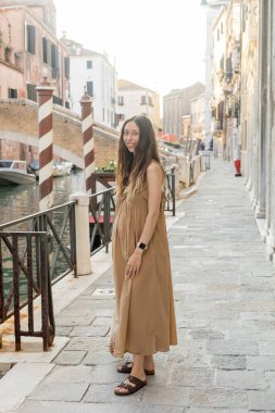 Cheerful pregnant woman with string bag looking at camera on street in Venice 