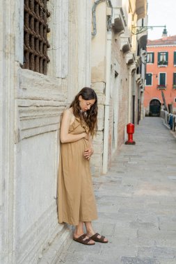 Young pregnant woman in dress touching belly on urban street in Italy 