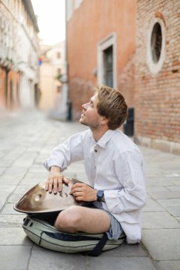 Side view of smiling musician playing handpan on urban street in Venice 