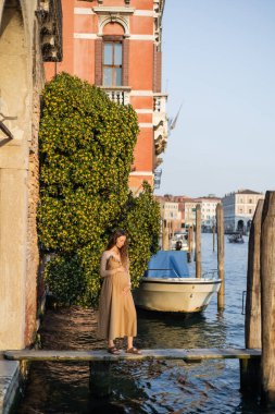 Pregnant woman in dress touching belly on wooden pier in Venice 