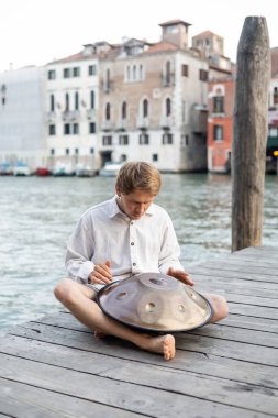 Barefoot musician playing hang drum on pier near river in Venice  clipart