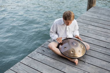 Overhead view of man playing hang drum on wooden pier above river in Venice 