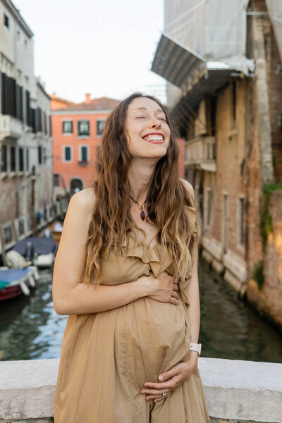 Happy pregnant woman touching belly in blurred Venice 