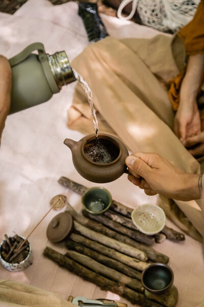 Cropped view of man pouring water in teapot near blurred tea bowls and wife in forest 