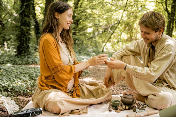 Smiling pregnant woman holding bowl near husband during tea ceremony in forest 