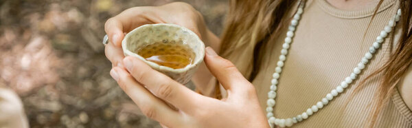 Cropped view of woman holding bowl with tea in forest, banner 