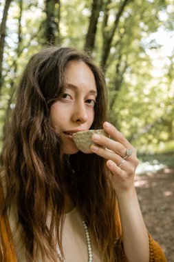 Young brunette woman drinking tea from bowl in forest 