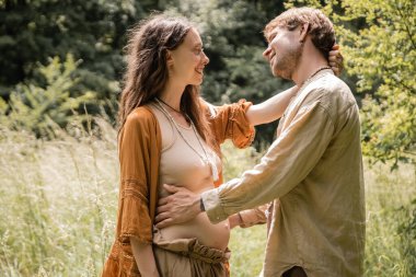 Side view of cheerful pregnant woman hugging husband in forest 