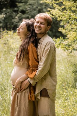 Smiling man hugging pregnant wife in summer forest 