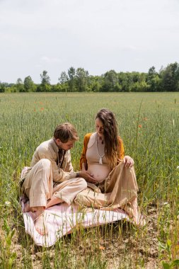 Man touching belly of pregnant wife on blanket in field 