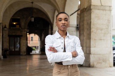 Kısa saçlı Afro-Amerikan turist kollarını kavuşturup Treviso 'daki bulanık şehir sokağındaki kameraya bakıyor.