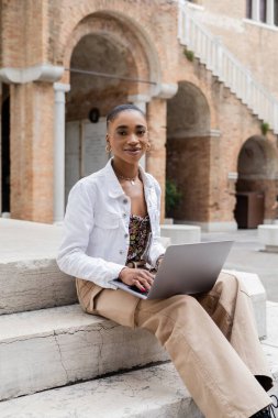 Laptop kullanan ve İtalya 'daki merdivenlerde kameraya bakan Afro-Amerikan blog yazarı.